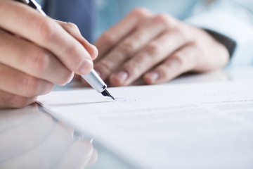 Zwei Hände, eine hält einen Stift und unterschreibt ein Dokument.