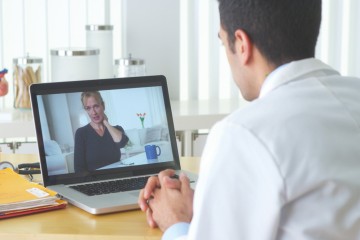 Ein Arzt sitzt am Schreibtisch und redet mit einer Patientin, die per Videosprechstunde zugeschaltet ist. 