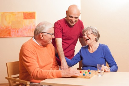Zwei Pflegebedürftige sitzen am Tisch und spielen. Ein Pfleger unterhält sich mit ihnen.