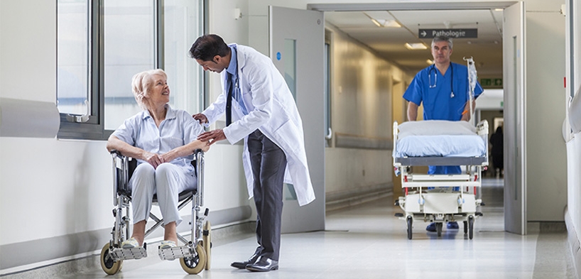 Krankenhausflur, eine Patientin im Rollstuhl mit einem Arzt im Gespräch, ein Arzt schiebt ein leeres Krankenhausbett