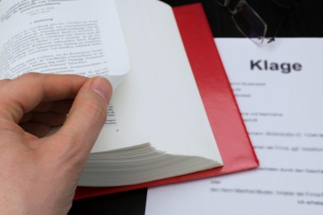 Die Finger der linken Hand blättern die Seite eines Gesetzbuches um. Darunter liegt ein Schriftstück mit dem Wort "Klage".
